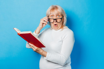 An old woman in glasses with a surprised face is reading a book on a blue background. Concept old...