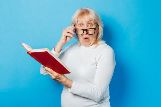 An old woman in glasses with a surprised face is reading a book on a blue background. Concept old lady reads books, education, book club.