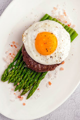 Beefsteak from minced beef with fried eggs and fresh green asparagus. Ketogenic food. Large portion on a white plate. Close up and vertical view.
