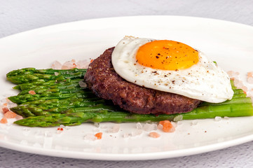 Beefsteak from minced beef with fried eggs and fresh green asparagus. Ketogenic food. Large portion on a white plate. Close up and horizontal view.