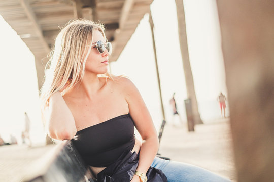Attractive blonde beautiful caucasian girl sitting on a bench with sunglasses and enjoying the outdoor urban vacation lifestyle in a sunny day - defocused people on the background