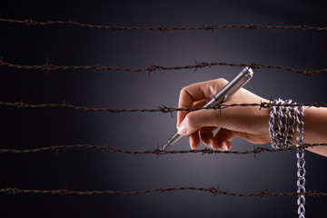 Woman hand with pen tied with chain and rusty sharp bare wire, depicting the idea of freedom of the press or expression. World press freedom and international human rights day concept. - Powered by Adobe