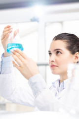 Serious thoughtful young female chemist in lab coat holding flask up while checking chemical liquid...