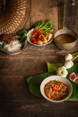 Thai Northern Style Pork and Tomato Chili Relish, nam prik ong in white bowl on wood table there are side dishes of fresh vegetables, cooked rice, water bowl and flower placed around.