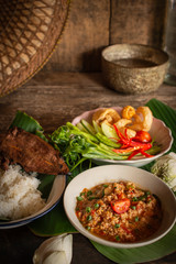 Thai Northern Style Pork and Tomato Chili Relish, nam prik ong in white bowl on wood table there are side dishes of fresh vegetables, cooked rice, water bowl and flower placed around.