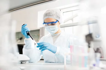 Foto op Plexiglas Ernstige geconcentreerde vrouwelijke microbioloog in steriele kleding en veiligheidsbril die aan tafel zit en reagens in petrischaal laat vallen terwijl hij onderzoek doet in het laboratorium © Seventyfour