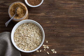 Raw oatmeal in a white bowl with peanut butter and cocoa. ingredients for a delicious chocolate porridge. healthy vegan breakfast on a wooden background