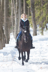 Fototapeta premium A winter forest. A young woman in warm jacket riding a black horse on snowy ground