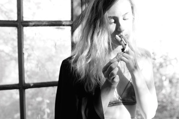 young blonde woman in bra smokes a cigarette by the window