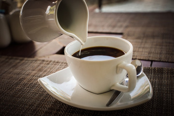Pouring milk into a white cup of hot black coffee