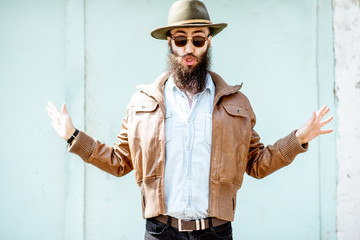 Portrait of a stylish bearded man dressed in jacket and hat on the light turquoise background outdoors