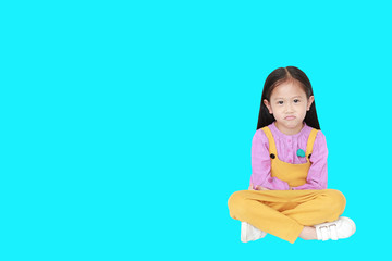 Portrait of angry little Asian child girl in pink-yellow pink-yellow dungarees sitting isolated on cyan background with copy space.