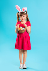 Little girl with basket of Easter eggs and bunny ears on color background