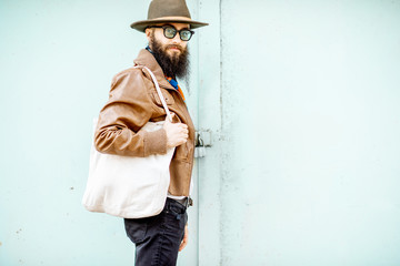 Portrait of a stylish man dressed in jacket, hat and scarf standing with bag on the turquoise background outdoors