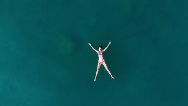 Young woman lie on back at sea waters, rest after swimming. Aerial shot, camera spin and fly up, reveal empty surface around still figure. Seabottom seen through crystal water of Aegean Sea