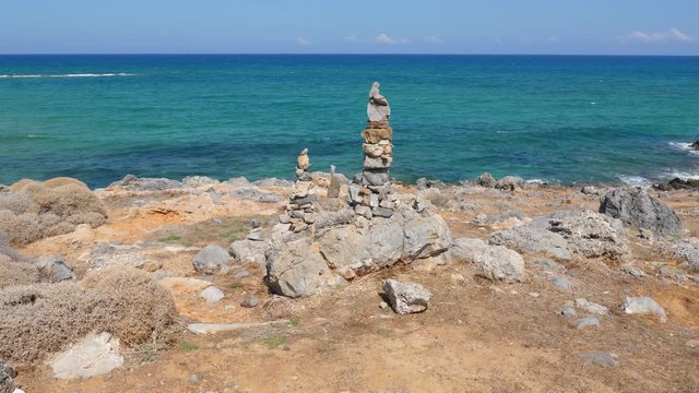 Stones stacked in small columns at sea shore, rough stony landscape around, hot sunny weather. Beautiful sea with turquoise water colour, seen vivid blue at horizon