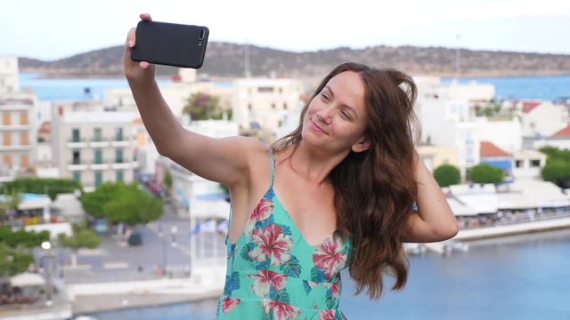 Attractive tourist woman take selfie pictures at Lake Voulismeni, half length portrait shot. She hold smartphone in hand, pose and correct hair, quickly look down. Blurred city buildings on background