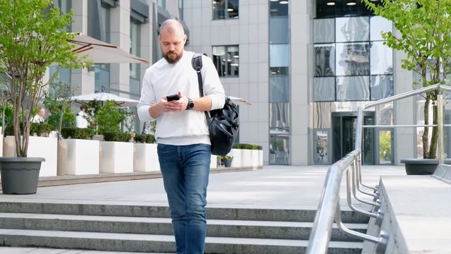 A man with a beard descends the steps of the office center, prints a message on his phone. Slow Motion