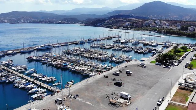 Aerial shot of large marina at Agios Nikolaos city, modern sailboats parked at safe area. Camera fly over empty square towards shelter for pleasure boats, bright sunny day at Crete