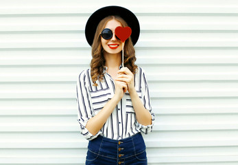 Portrait happy smiling young woman hiding her eye with red heart shaped lollipop in black round hat, white striped shirt on white wall background