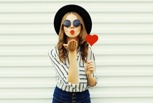 Portrait young woman blowing red lips sending sweet air kiss with red heart shaped lollipop in black round hat on white wall background