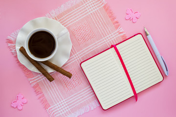 White porcelain cup with coffee, two coffee rolls on saucer with wavy edge and paper notebook with ball pen on a pink table napkin. Flat lay, copy space.