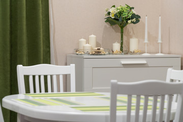 Kitchen with a wooden white oval table, white chairs, a green curtain, on the side of the candle in a candlestick and a flower