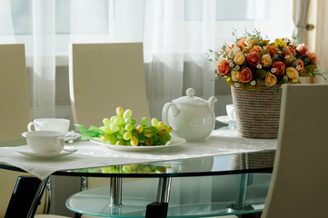 dining table setup with white porcelain crockery, cups and pot served for tea, basket with flowers, natural light from window. Modern interior, glass and metal surfaces, show room, restaurant, cafe
