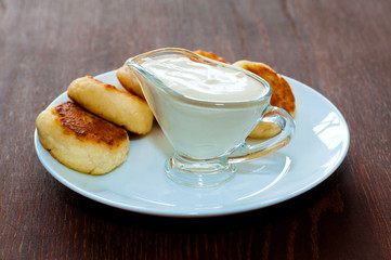 Fried cheese pancakes with sour cream on a white plate on a dark wooden table, background