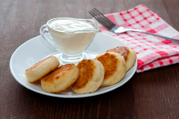 Cheese pancakes with sour cream on a white plate on a pink napkin with a fork, side view