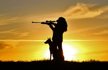 Fototapeta na wymiar Silhouette of a girl with a rifle at sunset with a dog, a breed of Belgian Shepherd Malinois dog