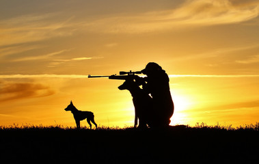 Silhouette of a girl with a rifle at sunset and two dogs, a breed of Belgian Shepherd dog Malinois and a miniature pinscher