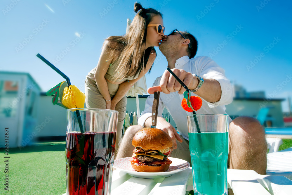 Wall mural young beautiful couple eating fast food by the pool, loving guy and girl eating Burger and smiling at each other