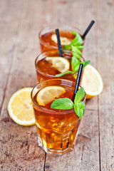 Traditional iced tea with lemon, mint leaves and ice cubes in three glasses on rustic wooden table.
