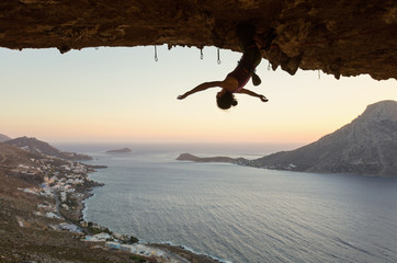 Female rock climber hanging upside down on challenging route in cave at sunset