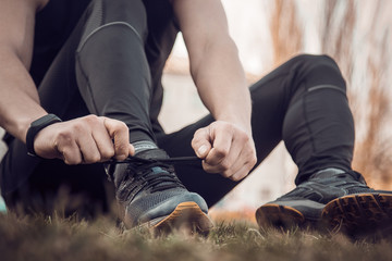 A young man in black clothes is tying the laces on the sneakers close up. fitness athlete sitting on the sports field on the grass. warm up body preparation for the training