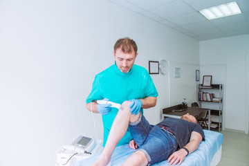 Young man receiving laser or magnet therapy massage on a knee to less pain. A chiropractor treats patient's knee-joint in medical office. Neurology, Osteopathy, chiropractic. Copy space.