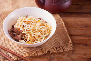 Asian noodles with beef, vegetables in bowl with chopsticks, rustic wooden background. Asian style dinner. Chinese Japanese noodles