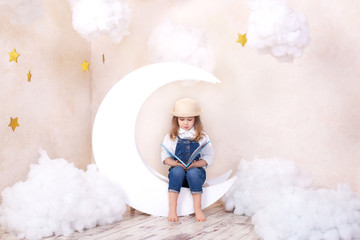 Little cute girl sitting on the moon with clouds and stars with a book in her hands and reading. The girl is learning to read. Reading useful hobby.  Kid cute bookworm. Pleasant time in cozy bedroom. 