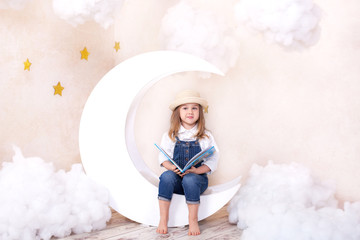 Little cute girl sitting on the moon with clouds and stars with a book in her hands and reading. The girl is learning to read. Reading useful hobby.  Kid cute bookworm. Pleasant time in cozy bedroom. 