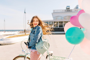 Happy smiling girl posing with bunch of helium balloons and white bicycle on the embankment while...