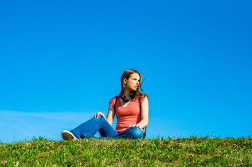 Naklejka na ściany i meble young teenager brunette girl in coral t-shirt with long hair sit on the grass on sky background with copy space