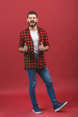 Full length portrait of a cheerful excited man in casual standing and showing thumbs up gesture with two hands isolated over red background.