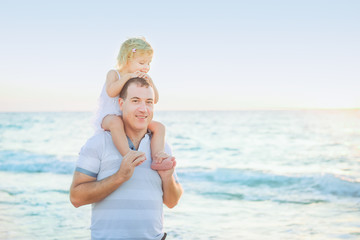 Portrait of young dad and his cute daughter sitting on his shoulders walking and having fun on the beach. Happy family at vacation. Sunset backlight. Soft selective focus. Copy space.