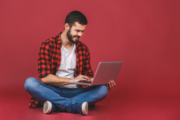 Portrait of a happy young man in casual holding laptop computer while sitting on a floor isolated...