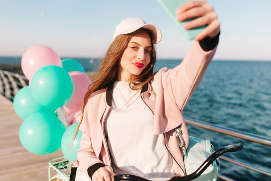 Tired girl with a stylish make-up is photographed at the sea background after morning bike ride along the pier. Gorgeous young long-haired woman in pink cap making selfie at the ocean in her birthday