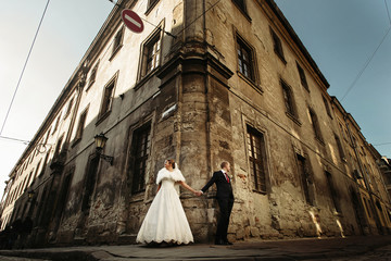 happy gorgeous bride and stylish groom holding hands and looking at different sides on background of old building