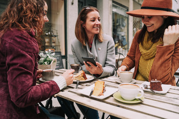 Three young women are talking animatedly while drinking coffee and eating some cake on a terrace of a bar. They are enjoying their company in a sunny day. Selective focus. Lifestyle