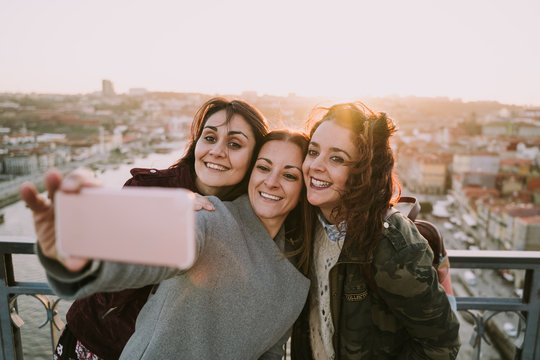 Three beautiful friends are on the Porto bridge sightseeing and taking a picture with a cell phone. The three girls are smiling happily. Lifestyle