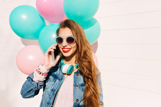Happy birthday girl with a hollywood smile accepts congratulations by smartphone holding bunch of balloons. Adorable brunette young woman wearing denim jacket planning a surprise party for best friend